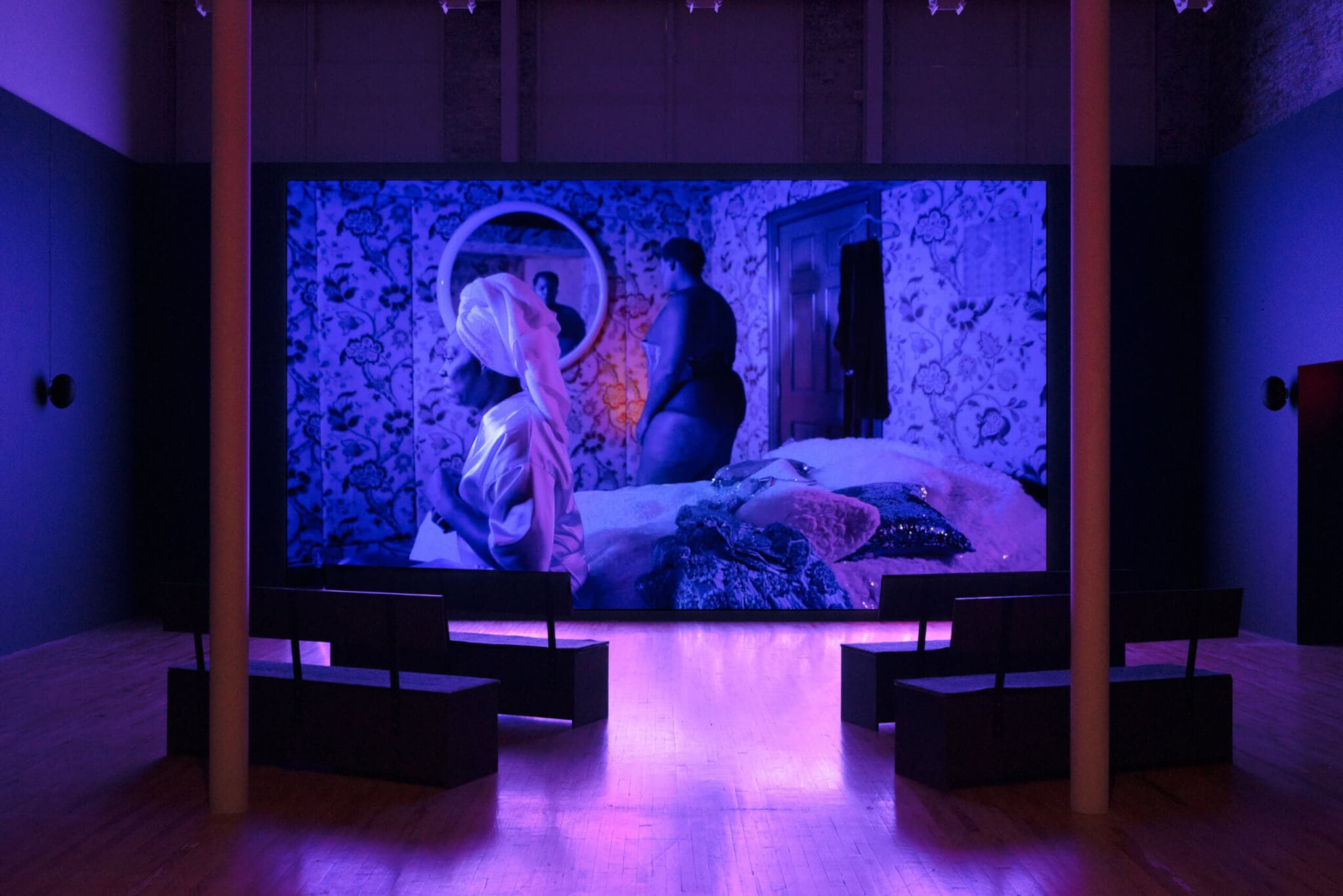 A video installation displays a figure looking into a mirror, while the room is lit by purple and blue light.